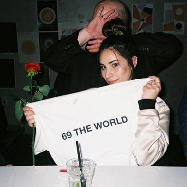 Girl holding shirt that says '69 the world' as man behind her covers his face